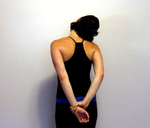 Woman in black tank top with hands clasped behind her back against white wall.